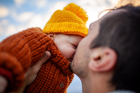 Happy man kissing his little baby girl walking outdoors. Father holding daughter wearing fall brown sweater and yellow hatの写真素材