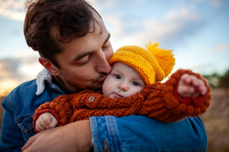 Portrait of young man kissing little baby girl walking outdoors. Father holds daughter wearing fall brown sweater and yellow hatの写真素材