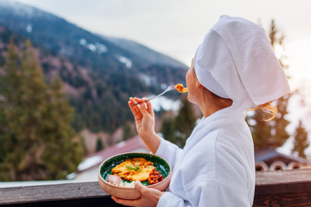 Woman ordered breakfast in hotel room eating food on balcony wearing robe enjoying winter mountains landscape. Vacationの写真素材