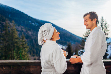 Young couple man and woman drinking coffee on hotel balcony enjoying winter mountains view wearing white robes. Spaceの写真素材