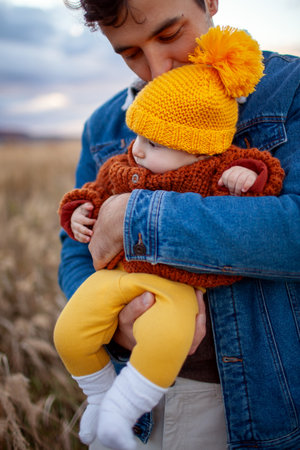 Happy man kissing his little baby girl walking outdoors. Father holding infant daughter wearing fall brown sweater and yellow hatの写真素材