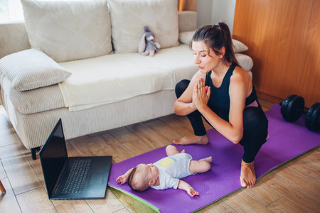 Postpartum recovery workout at home. Young woman exercising together with little baby girl on yoga mat. Squatting with infant using laptop online courseの写真素材