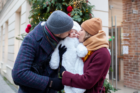 Portrait of parents kissing their baby girl on city street decorated for Christmas. Family enjoys...