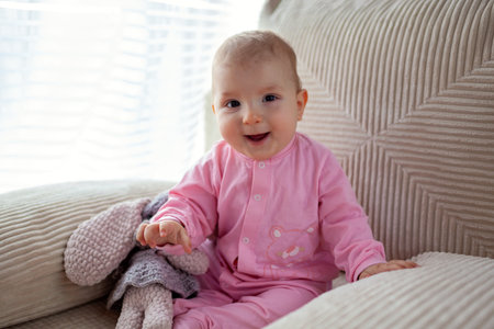 Happy infant baby girl playing on couch at home wearing pink bodysuit. Cute child smiling.の写真素材