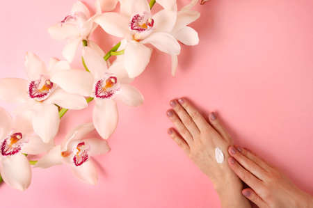 Woman applyiing cosmetic cream on her hands on rose background with orhid flower. Home spa, beauty and treatment concept. Flat lay, top view.の写真素材