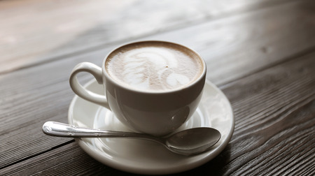 Cup of tasty latte is standing on the wooden textured table. There is nice picture is painted on the top of the latte. Latte is in the white big cup.の写真素材