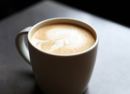Cup of tasty latte is standing on the grey textured table. There is nice picture is painted on the top of the latte. Latte is in the white big cup.の写真素材