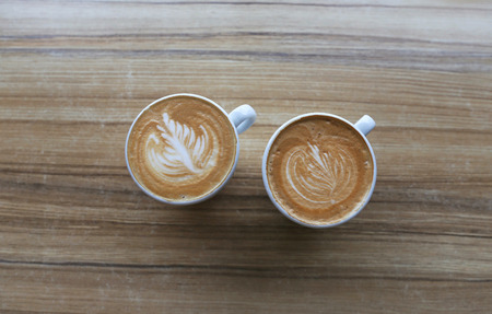 Two Cups of tasty cappuccinno are standing on the wooden textured table.  Cups are having some painting on the top of coffeeの写真素材