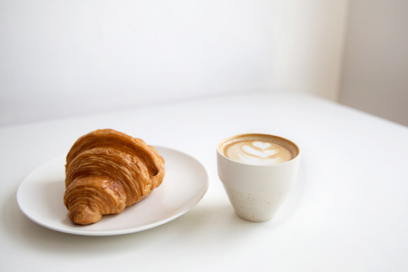 Cup of cappuccino coffee and croissant on white tableの写真素材