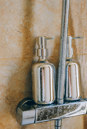 Close-up of sleek, silver soap dispensers against a marble bathroom wall, with a modern shower tap...