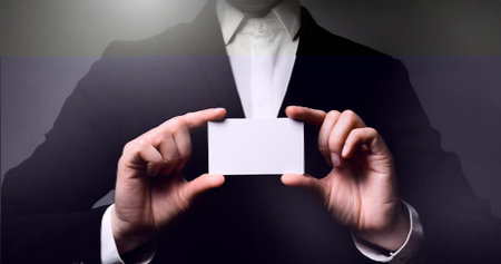 Businessman holding a blank business card in front of a dark backgroundの素材