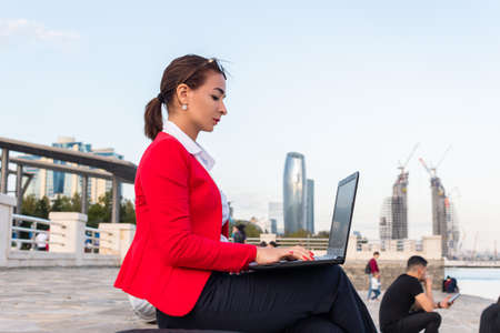 a businesswoman in a red suit sits on the seaside parkway with a laptop. Freelancer.の写真素材