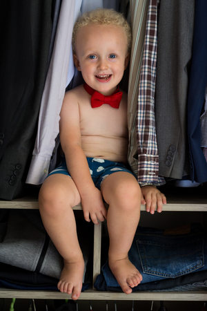 Cute and handsome little boy with curly blond hair wearing underpants and red bow tie, seating and smiling in dad's wardrobe. Little gentleman is getting ready. Happy father's day concept.の写真素材