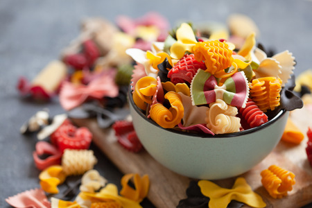 Various colourful uncooked pasta in blue bowl on wooden board over stone background, close upの写真素材