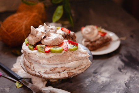 Vegan tropical aquafaba cake with kiwi, strawberry and coconut cream on dark table with ingredients and flowers, close up. Concept of vegetarian dessert for celebrationの写真素材