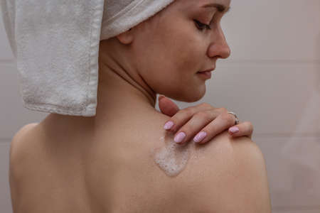 Beautiful young woman with a white towel on her head standing in the bathroom and applying shower foam to her shoulder.の写真素材