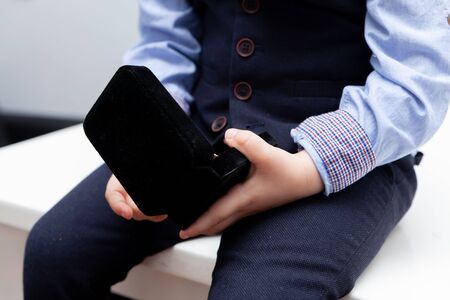 A boy in a shirt, bow tie, vest and trousers on a white background is holding a jewelry box. Stylish...