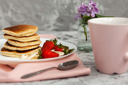 A stack of pancakes with a pink tea mug and fresh strawberries on a white plate on a light...