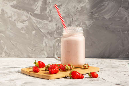 Strawberry smoothie in a glass jar with a red-striped tube. There are strawberries on a wooden Board and a strawberry smoothie next to them. Concept of healthy food. On a light concrete backgroundの写真素材