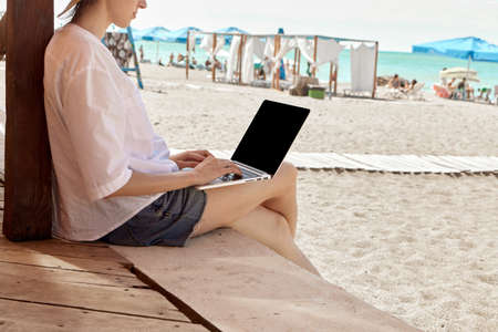 Young woman using laptop computer on a beach. Freelance work concept. Pretty young woman using laptop in cafe on tropical beach in outdoor cafe terrace with sea view. Work and travelの写真素材