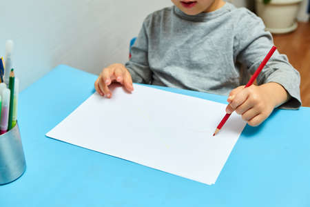 Cute little boy with blond hair draws colored pencils at home. Draws at the blue table. Close up of child's hands drawing at white paperの写真素材