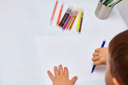 Cute little boy with blond hair draws colored pencils at home. Draws at the white table. Close up of...