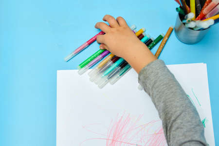 Cute little boy with blond hair draws colored pencils at home. Draws at the blue table. Close up of child's hands drawing at white paperの写真素材
