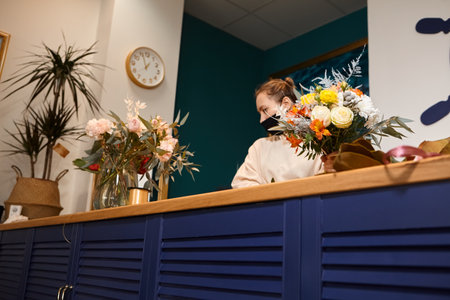 Florist works with colors. Flower seller chooses flowers for future bouquet. Flowers shop worker in a mask standing in flower shop and checking flowers in glass vase.の写真素材