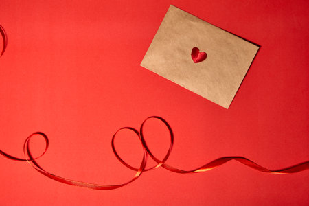 A red heart lies on a brown envelope for Valentine's Day. Red backdrop with envelope and red heart. Love letter or message concept. Flat-lay, top view. Copy space for your text.の写真素材