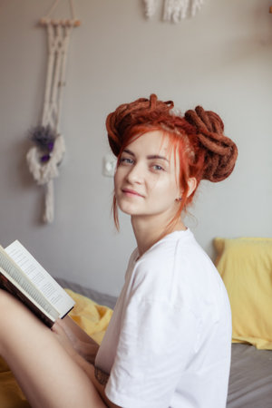 Very pretty redhead girl with dreads on bed in bedroom in the morning. Young woman reading a book sitting in bed. A feminine, gentle girl.の写真素材