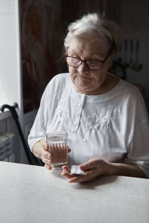 Sad old woman taking pills, health problems in old age, expensive medications. An elderly woman's hands unpacking several pills for taking medication. Grandma takes tablet and drinks a glass of waterの写真素材