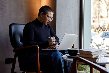A young girl with glasses, with short hair is sitting in a cafe and working on a laptop. The girl looks at her laptop and makes notes in a notebook. The concept of distance learningの写真素材