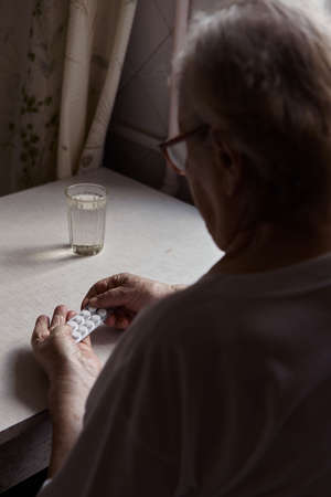 Sad old woman taking pills, health problems in old age, expensive medications. An elderly woman's hands unpacking several pills for taking medication. Grandma takes tablet and drinks a glass of waterの写真素材