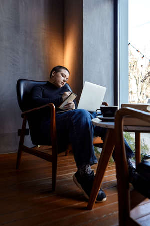 A young girl with glasses, with short hair is sitting in a cafe and working on a laptop. The girl looks at her laptop and makes notes in a notebook. The concept of distance learningの写真素材