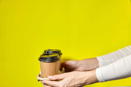 Hands holding two cups brown paper with black lid. Two coffee special offer or promo. Hands holding two cups on yellow screen background. Tea or Coffee to go. Brown paper cup with black lid.の写真素材