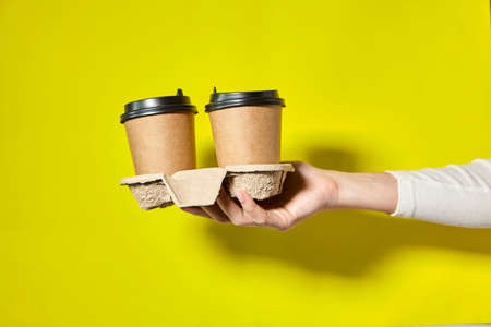 Hands holding two cups brown paper with black lid. Two coffee special offer or promo. Hands holding two cups on yellow screen background. Tea or Coffee to go. Brown paper cup with black lid.の写真素材