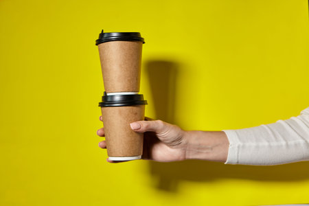 Hands holding two cups brown paper with black lid. Two coffee special offer or promo. Hands holding two cups on yellow screen background. Tea or Coffee to go. Brown paper cup with black lid.の写真素材