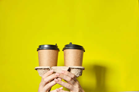 Hands holding two cups brown paper with black lid. Two coffee special offer or promo. Hands holding two cups on yellow screen background. Tea or Coffee to go. Brown paper cup with black lid.の写真素材
