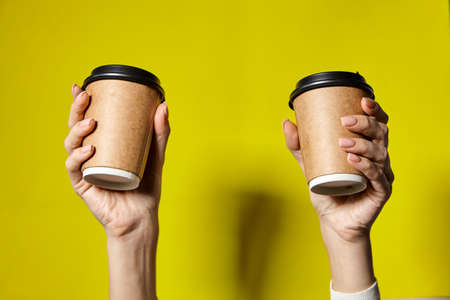 Hands holding two cups brown paper with black lid. Two coffee special offer or promo. Hands holding two cups on yellow screen background. Tea or Coffee to go. Brown paper cup with black lid.の写真素材