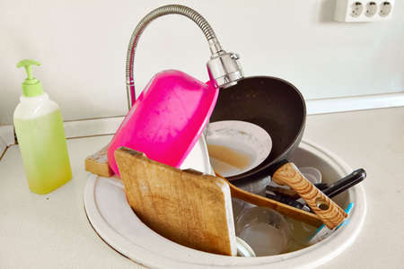 A woman in an apron and rubber gloves washes dirty dishes in the sink in her kitchen. Dirty dishes in a sink for washing up.の写真素材