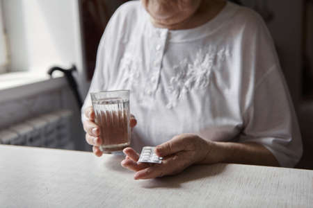 Sad old woman taking pills, health problems in old age, expensive medications. An elderly woman's hands unpacking several pills for taking medication. Grandma takes tablet and drinks a glass of waterの写真素材