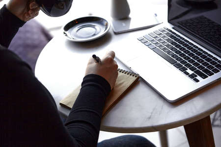 A young girl is sitting in a cafe, working on a laptop and making notes in a notebook. Close - up photo of hands. The concept of freelancing and remote work or training.の写真素材