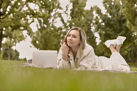 The woman is lying down using a laptop in the park. Smiling young student woman is using a laptop laying on grass.の写真素材