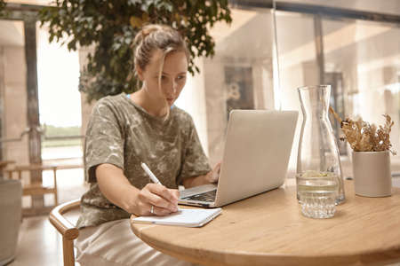 Portrait beautiful young woman sitting at table with laptop in cafe. Learning online, typing, search for information, notes of the synopsis.の写真素材