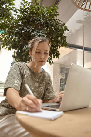 Portrait beautiful young woman sitting at table with laptop in cafe. Learning online, typing, search for information, notes of the synopsis.の写真素材