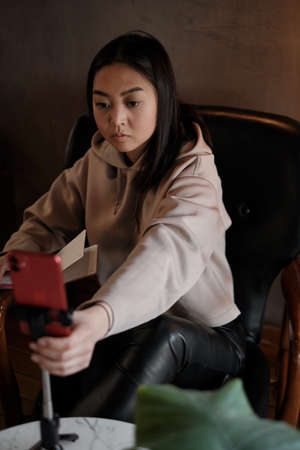 Distance learning. Asian female student sitting in cafe, looking at phone and writing in writing notebook.の写真素材
