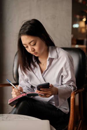 Asian female sitting in cafe, holding phone and pencil writing notebookの写真素材