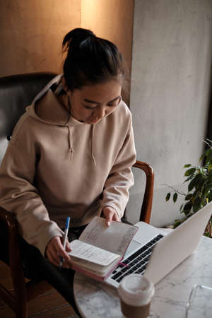 Distance learning. Asian female student sitting in cafe, looking at phone and writing in writing notebook.の写真素材