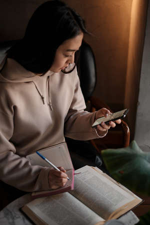 Shot of young asian female student sitting at table and writing on notebook. Young female student...