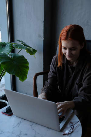 a red-haired freelance woman with glasses works remotely on a laptop in a cafe.xの写真素材